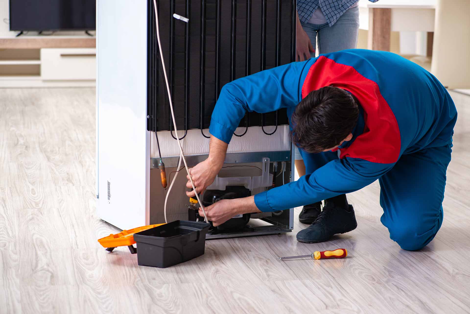 Technician kneels behind a refrigerator to repair the freezer